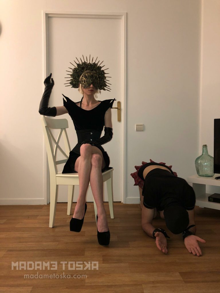 Madame Toska, dominatrice à Paris et son soumis. Elle est assise tandis qu'il se tient à quatre pates en position de soumission près d'elle avant de subir plusieurs minutes de gode-ceinture et travail anal en tout genre..